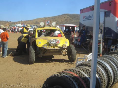 1990 Ford Bronco - yellow off-road race buggy with roof lights sits in a busy desert pit lined with spare tires and crew.