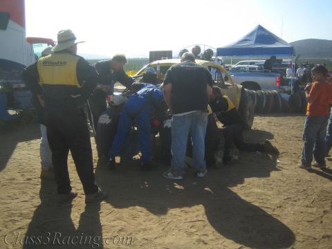 1990 Ford Bronco - crowd of crew members huddles around a yellow off-road race buggy in a sunlit, dusty desert pit area.