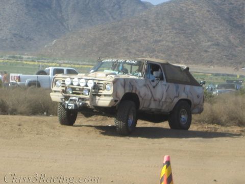 1990 Ford Bronco - camouflage-painted lifted Bronco-style SUV with a soft top crosses a dusty desert flat near a course cone.