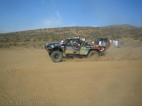 1990 Ford Bronco - black number 719 off-road race truck speeds across a desert flat, throwing up a heavy trail of dust.