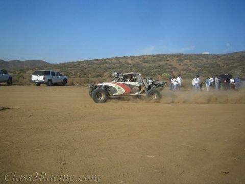 1990 Ford Bronco - off-road desert race buggy marked number 10 speeds across a dusty dirt course as spectators and trucks watch from the roadside.