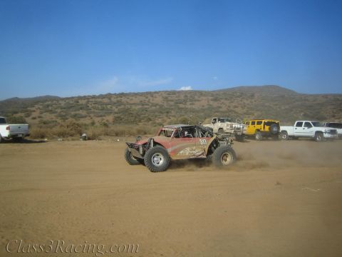 1990 Ford Bronco - red off-road race buggy numbered 511 races over a dusty desert track with parked chase vehicles in the background.