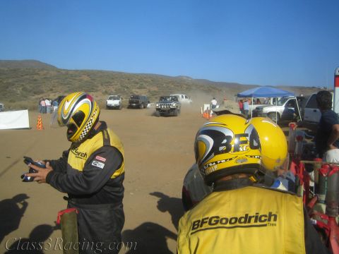 1990 Ford Bronco - Two pit crew members wearing yellow BFGoodrich jackets and helmets stand at a dusty desert race area as vehicles approach.