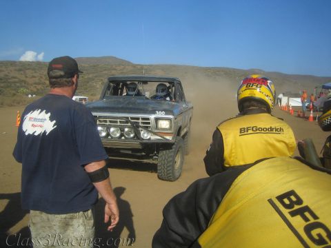 1990 Ford Bronco - black Ford Bronco off-road race truck numbered 300 rolls into a desert pit as crew members in BFGoodrich gear look on.