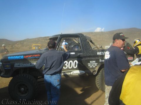 1990 Ford Bronco - black race truck numbered 300 covered in sponsor decals gets attention from crew during a desert pit stop.