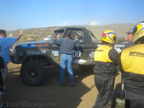 1990 Ford Bronco - Crew members in BFGoodrich suits service a black sponsor-covered off-road race truck during a desert pit stop.