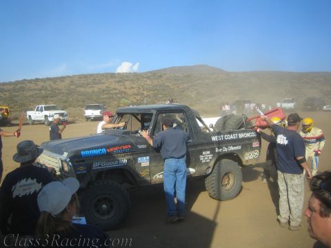 1990 Ford Bronco - black West Coast Broncos race truck numbered 300 is refueled and serviced by a crowd of crew at a dusty desert pit.