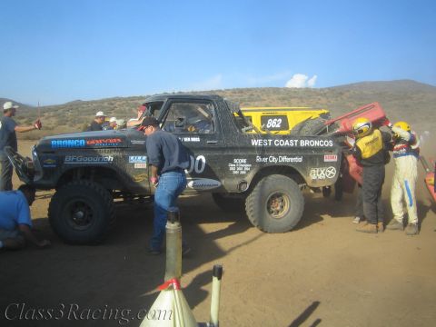 1990 Ford Bronco - black race truck numbered 300 sits beside a yellow truck numbered 862 as crews work in a dusty desert pit.