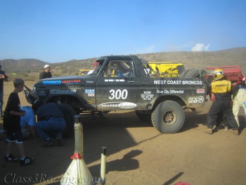 1990 Ford Bronco - Crew members service the black West Coast Broncos truck number 300, one kneeling at a front wheel, in a desert pit.