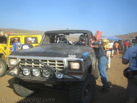 1990 Ford Bronco - black off-road race truck number 300 sits front-on with its multi-light bar as crew and spectators crowd the desert pit.