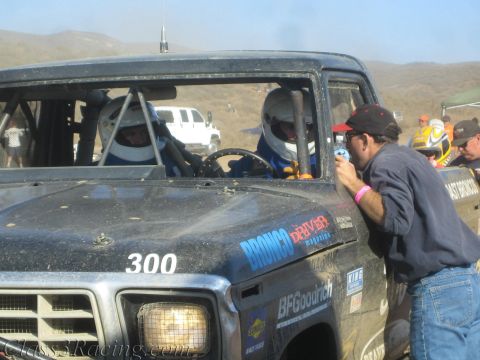 1990 Ford Bronco - Two helmeted drivers sit in the cab of black race truck number 300 while a crew member leans in at the window.