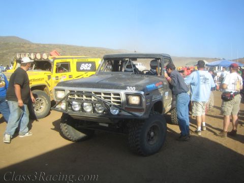 1990 Ford Bronco - black off-road race truck numbered 300 with a front light bar sits in a crowded desert pit beside a yellow truck.