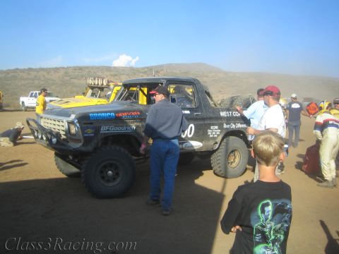 1990 Ford Bronco - black West Coast Broncos race truck is serviced by crew at a desert pit as a young boy in a black shirt watches.