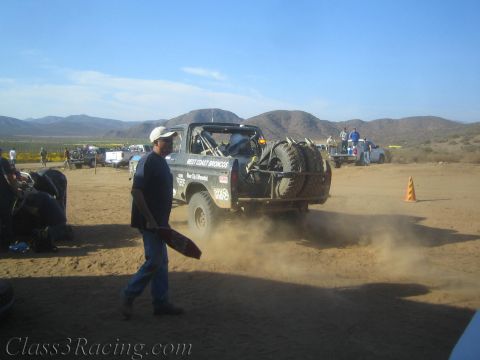 1990 Ford Bronco - black West Coast Broncos race truck pulls away in a cloud of dust from a desert pit, with mountains in the distance.