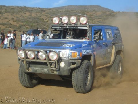 1990 Ford Bronco - blue Hummer H3 off-road race truck numbered 760 with a roof light bar charges through dust as spectators watch.