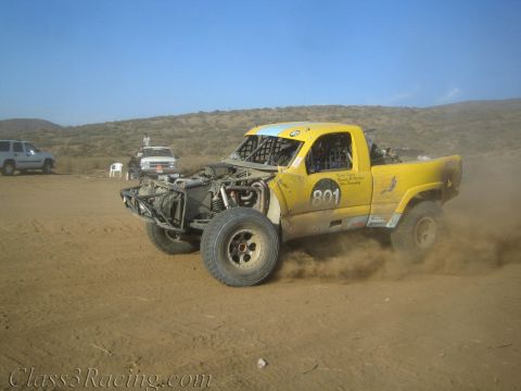 1990 Ford Bronco - yellow off-road race truck numbered 801 throws up dirt as it races across a desert track.