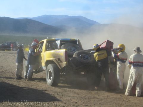 1990 Ford Bronco - Crew members handle red fuel jugs while servicing a yellow off-road race truck at a dusty desert pit framed by mountains.