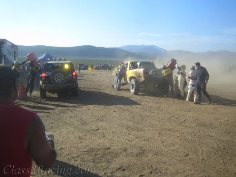 1990 Ford Bronco - Off-road race trucks and crew gather in a hazy, dust-filled desert pit area under morning light with mountains beyond.
