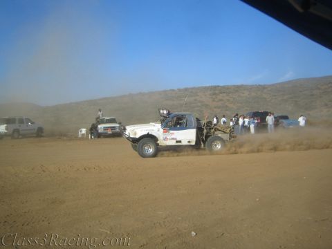 1990 Ford Bronco - white off-road race truck slides through dust on a desert course as spectators and chase trucks line the route.