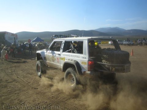 1990 Ford Bronco - white Howard Roofing off-road race SUV kicks up dust on a desert course with a spare tire on the rear and mountains behind.
