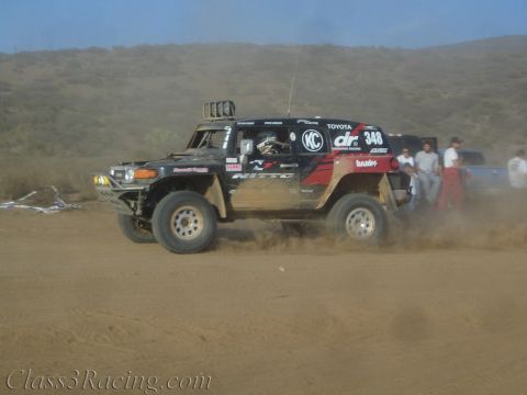 1990 Ford Bronco - black and red Toyota FJ-style off-road race truck numbered 348 catches air on a dusty desert course.