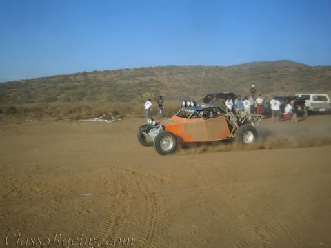 1990 Ford Bronco - orange open tube-frame off-road buggy speeds across a desert track as spectators and trucks watch from the background.