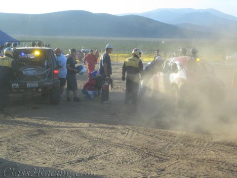 1990 Ford Bronco - Crew members and race vehicles crowd a dusty, backlit desert pit at dusk with green fields and mountains behind.