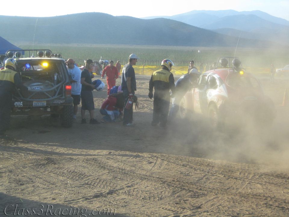 Race vehicles and crew gathered in a dusty, hazy desert pit at dusk near mountains
