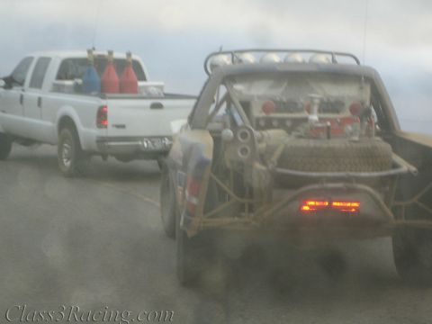 1990 Ford Bronco - tube-frame off-road race truck with glowing tail lights is towed behind a white pickup carrying fuel jugs in foggy weather.