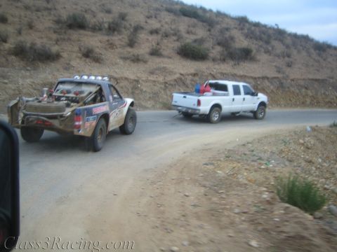 1990 Ford Bronco - sponsor-marked off-road race truck is towed by a white pickup truck along a winding dirt road through dry hills.