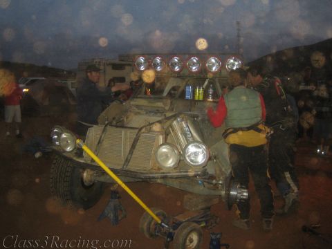 1990 Ford Bronco - stripped off-road race buggy sits on jack stands with a light bar and big front headlights as crew work at a dim desert pit.