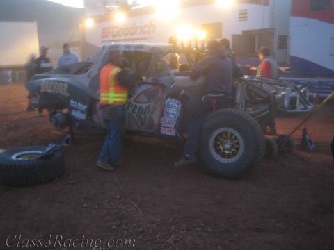 1990 Ford Bronco - Crew members surround a muddy off-road race truck with spare tires nearby under a lit BFGoodrich banner at night.