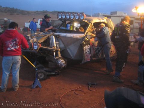 1990 Ford Bronco - crew works on a battered tan off-road race car raised on a jack with a roof light bar in a dusty night pit.