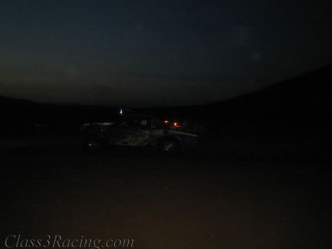 1990 Ford Bronco - faint off-road race truck shows its lights against a dark dusk landscape on the racecourse.