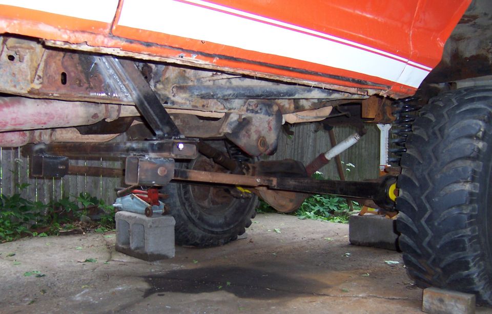 Vehicle undercarriage with custom suspension parts and large tires. It's on cinder blocks with a jack visible