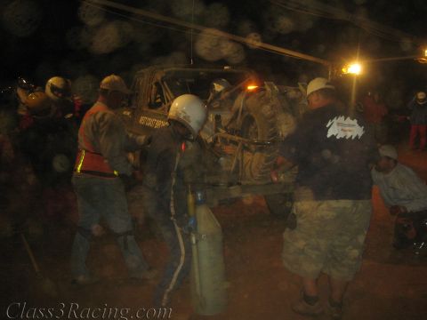 1990 Ford Bronco - Helmeted crew gather around a dim off-road race truck at night, one holding a gas bottle, under string lights.