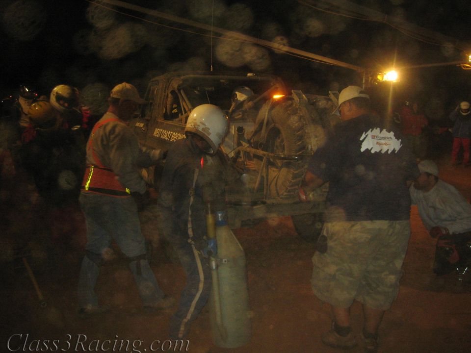 Crew in helmets around a race truck at night with oxygen-style bottle