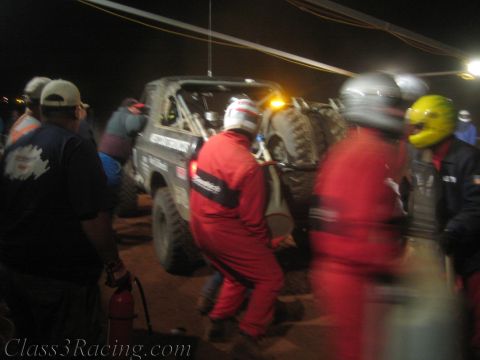 1990 Ford Bronco - Pit crew in red driving suits and helmets rush around a lifted off-road race truck at night with a fire extinguisher ready.