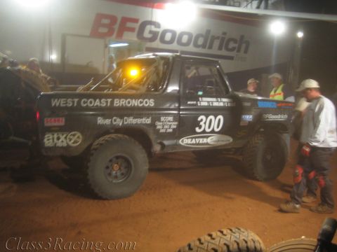 1990 Ford Bronco - black off-road race truck marked West Coast Broncos number 300 sits under a lit BFGoodrich banner with crew alongside.