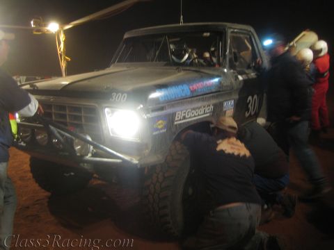 1990 Ford Bronco - black off-road Bronco race truck number 300 shines its headlights as crew kneel to change a front tire at night.