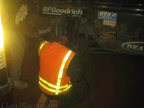 1990 Ford Bronco - crew member in an orange reflective vest crouches by a muddy tire beneath a black off-road race truck plastered with stickers.