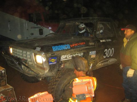 1990 Ford Bronco - black off-road race truck number 300 with a helmeted driver sits in a dusty night pit as crew work the front wheel.