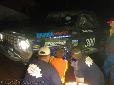 1990 Ford Bronco - Crew members in vests kneel at the lit front of a black off-road race truck number 300 working the wheel at night.