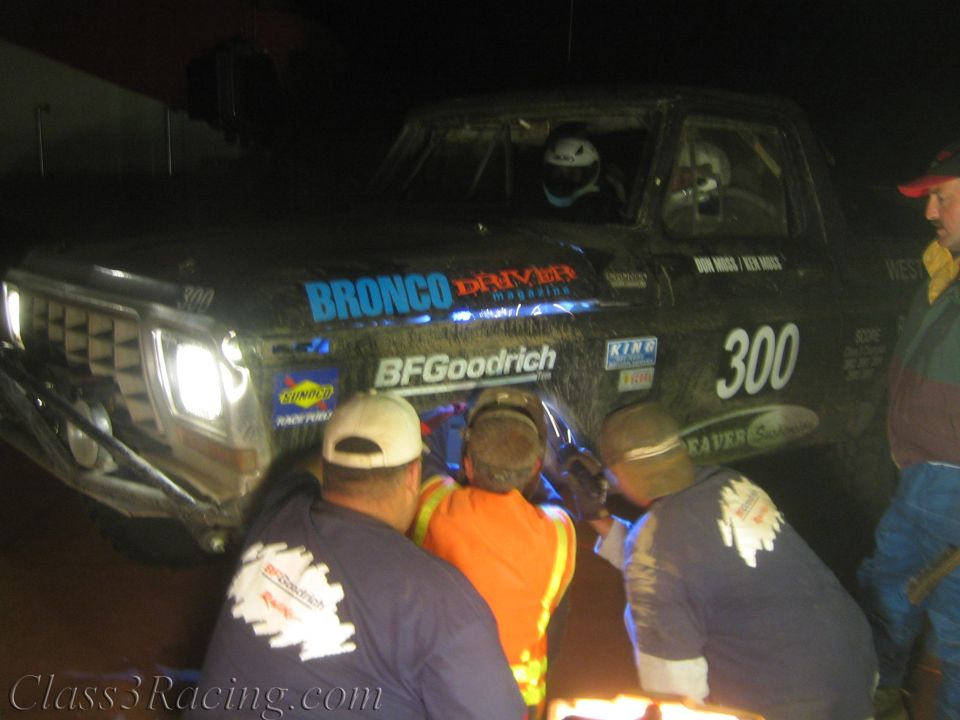 Crew kneeling at the front wheel of a black race truck with headlights on