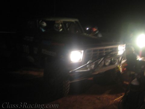 1990 Ford Bronco - off-road race truck glares its headlights into the camera amid darkness and bright pit lighting at night.