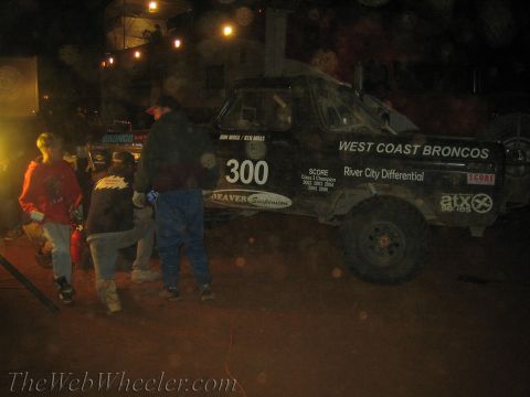 1990 Ford Bronco - black West Coast Broncos number 300 race truck sits in a hazy night pit as several crew gather beside it.