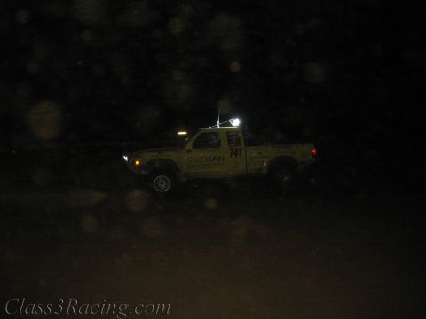 1990 Ford Bronco - yellow off-road race truck lettered Guzman number 741 runs the dark course with its lights and roof light glowing.