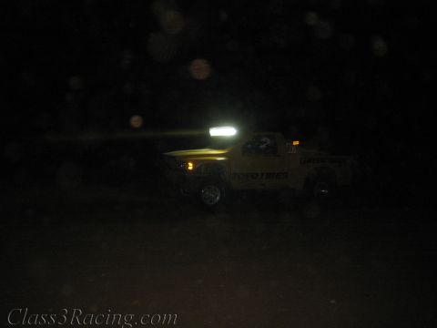 1990 Ford Bronco - yellow off-road race truck marked Toyo Tires beams a roof light bar while running on a dark night course.