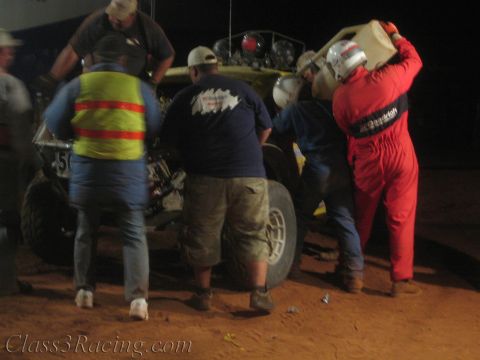 1990 Ford Bronco - Crew surround a lifted yellow off-road race buggy at night as one in a red suit carries a fuel jug on his shoulder.