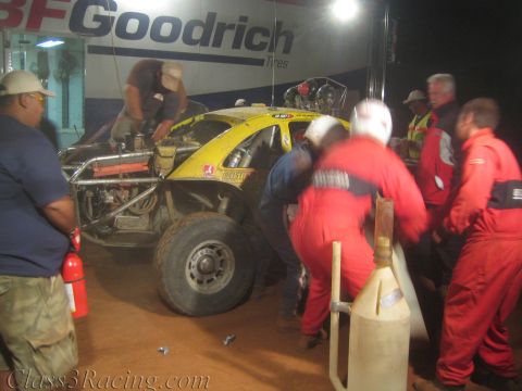 1990 Ford Bronco - Crew in red suits service an open yellow off-road race buggy under a lit BFGoodrich banner, with a fire extinguisher and fuel jug nearby.